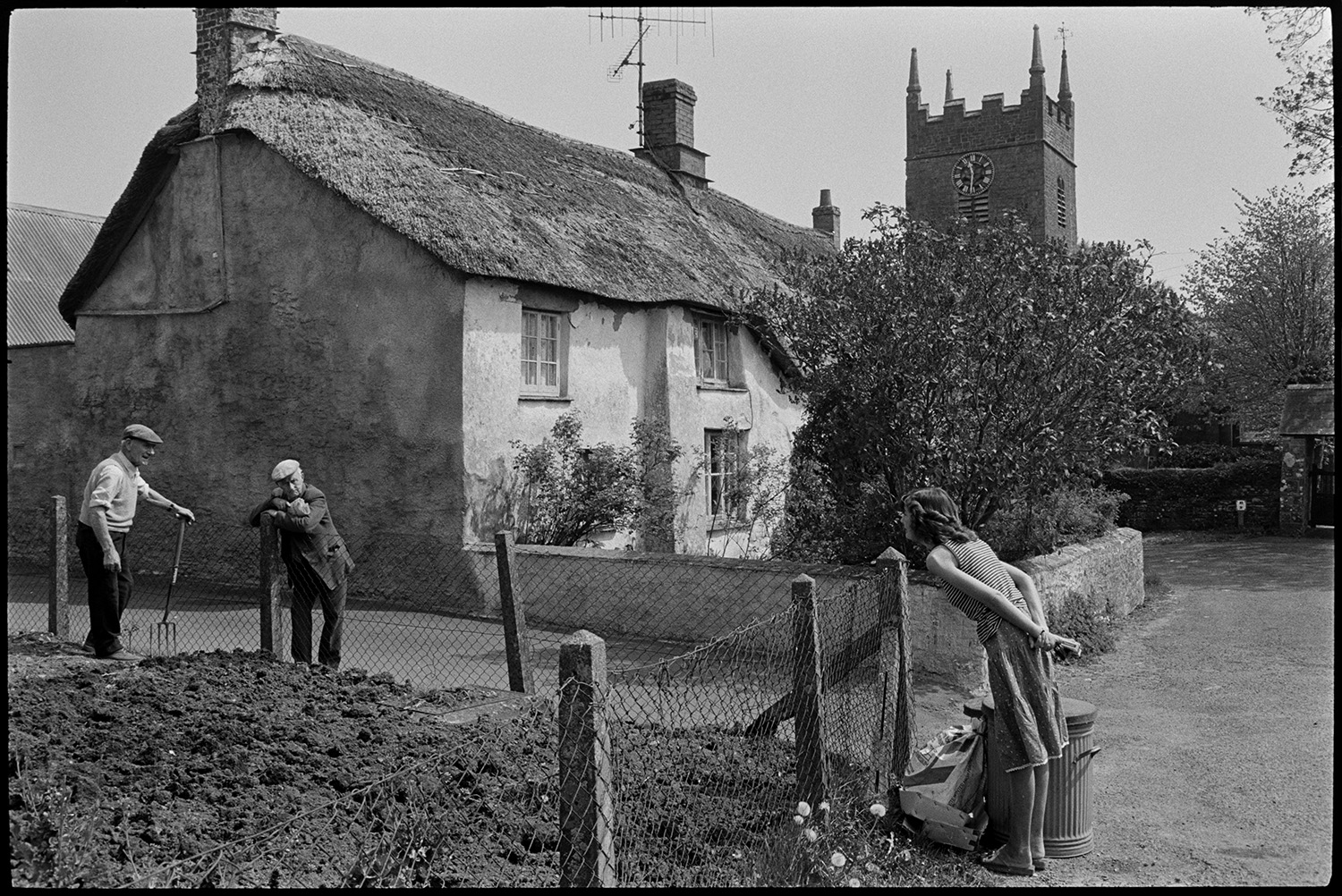 Street scenes, house with porch, thatched farmhouse and door, people chatting, church tower. 
[A woman and man leaning over a garden fence to talk to a man gardening, in Northlew. The woman is stood by rubbish bins. A thatched cottage and the church tower can be seen in the background.]