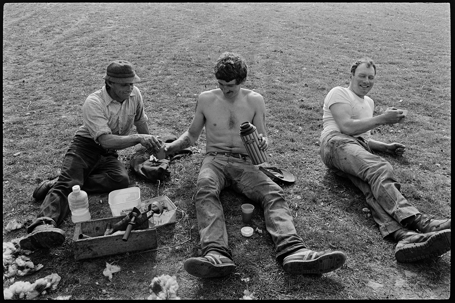 Sheepshearers tea break in field. 
[Three men having a break from sheep shearing in a field at Brendon Barton. They are sat on the grass and are pouring drinks from a thermos flask.]