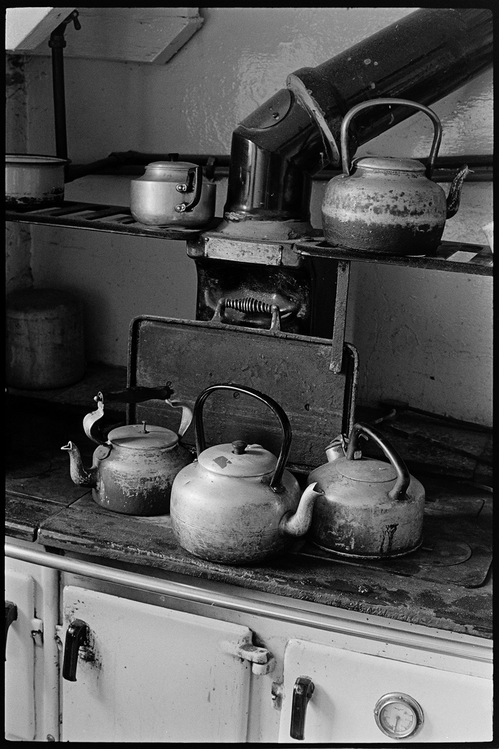 Rayburn stove with kettles on hotplate. 
[Three kettles on the hotplate of a rayburn stove in a kitchen in a house in Brendon Barton. Other kettles are positioned on a rack above the stove.]