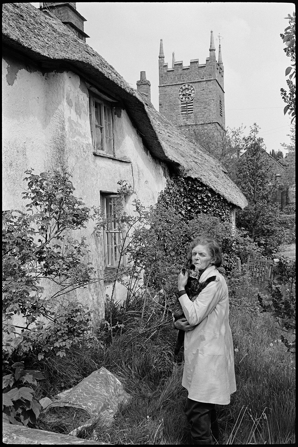 House with porch, people chatting, slate roofed workshop or forge? Church tower. 
[A woman holding a cat stood outside a thatched cottage in Northlew. The church tower can be seen in the background.]