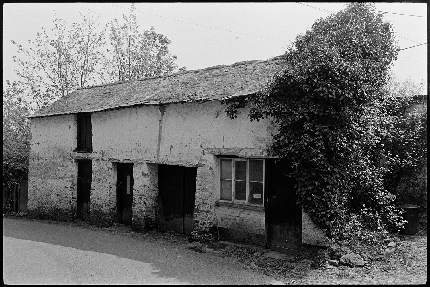 House with porch, people chatting, slate roofed workshop or forge? Church tower. 
[A slate roofed building overgrown with ivy at one end in Northlew. It was possibly a workshop or a forge.]