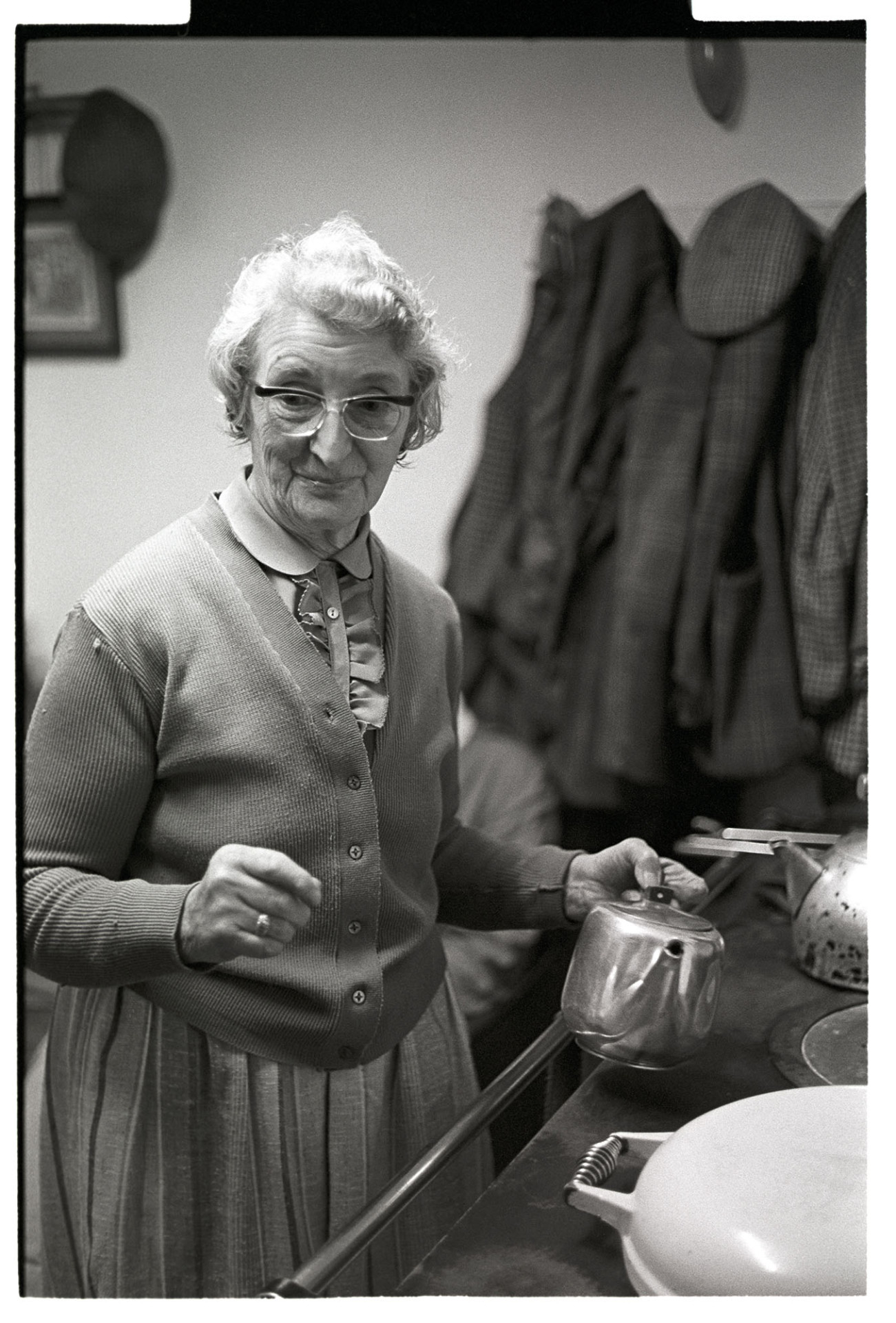 Woman standing in front of stove pouring tea. 
[A woman stood by a stove holding a teapot at an Exmoor farm. Coats and hats are hung up on a wall in the background.]