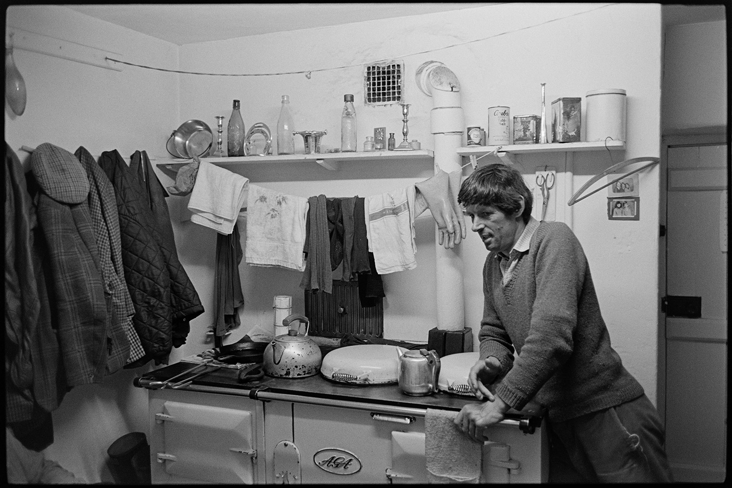 Man leaning on stove, kettle, teapot, cloths drying, mantelpiece. 
[A man leaning on a Aga at Exmoor Farm. Kettles are on the stove and tea towels are hung up to dry above the stove. Coats are hung up on the wall and a mantelpiece above the stove has various ornaments, including candlesticks and tins, on display.]