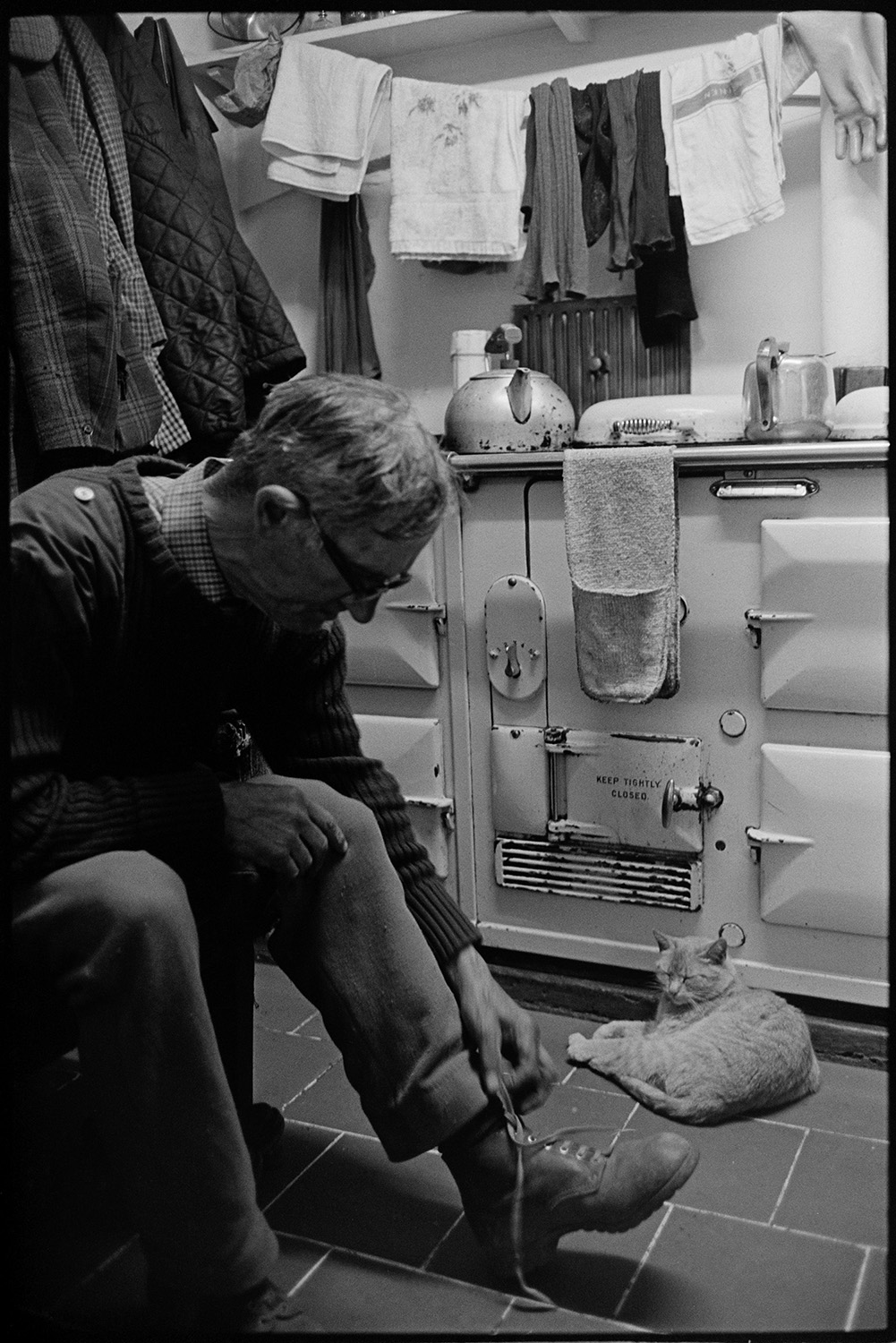 Man leaning on stove, kettle, teapot, cloths drying, mantelpiece. 
[A man sat on a chair untying his boot laces, by an Aga at Exmoor Farm. A kettle and pair of oven gloves are on the stove and a cat is sat in front of the Aga. Coats are hung up on the wall in the background.]