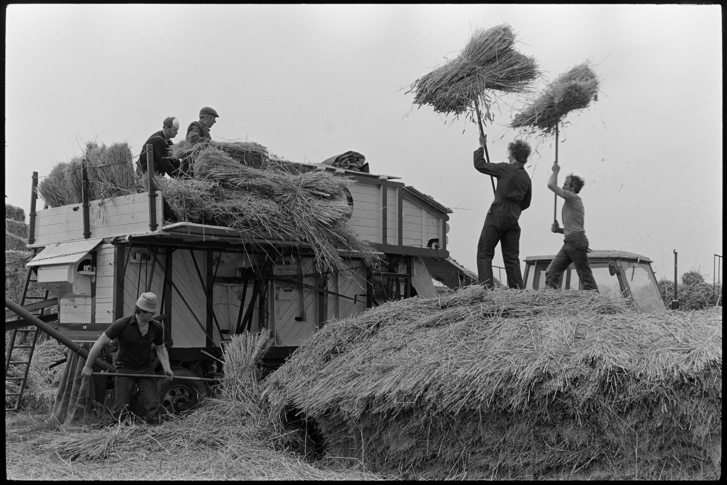 Reed comber at work, pile of ropes and spars. 
[Men dismantling a wheat rick and loading the reed into a reed comber, using pitchforks, in a field at Westacott Barton, Riddlecombe. Two of the men are holding bundles of reed above their heads on pitchforks.]