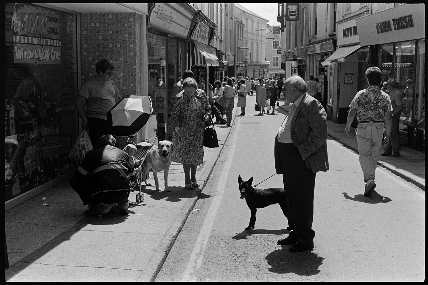 Street scenes with shoppers and passers by. 
[Men and women shopping in Bideford High Street. Two people are walking dogs and another person is checking on a child in a pushchair. Shop fronts are visible, including Farm Fresh and R Mackenzie Dye.]