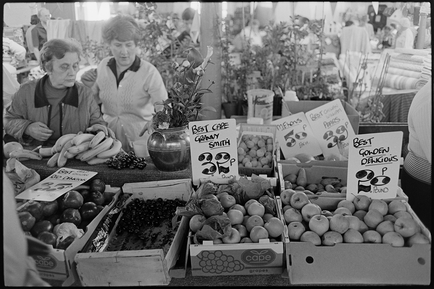 Fruit stall at Pannier Market showing price tags. 
[Two women looking at a fruit stall at Bideford Pannier Market selling apples, cherries, melons, bananas and pears. The prices of the fruit is advertised.]