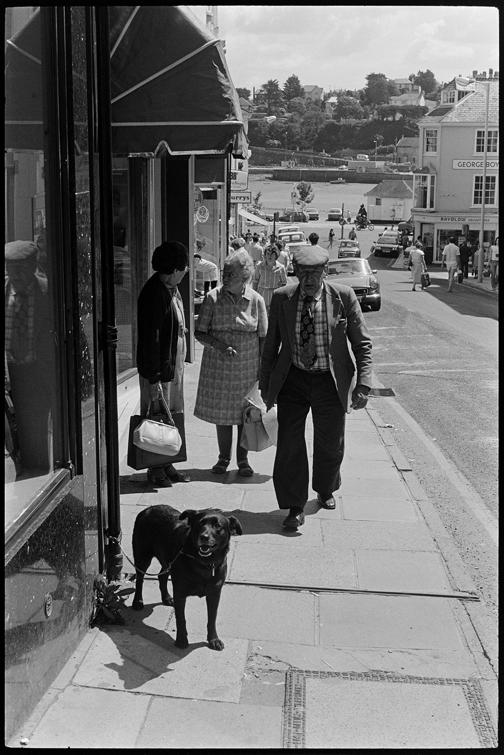 Street scenes with shoppers and passers by. 
[Men and women chatting and walking along Bideford High Street past shop fronts. A dog is tethered to a drainpipe outside one of the shops with an awning.]