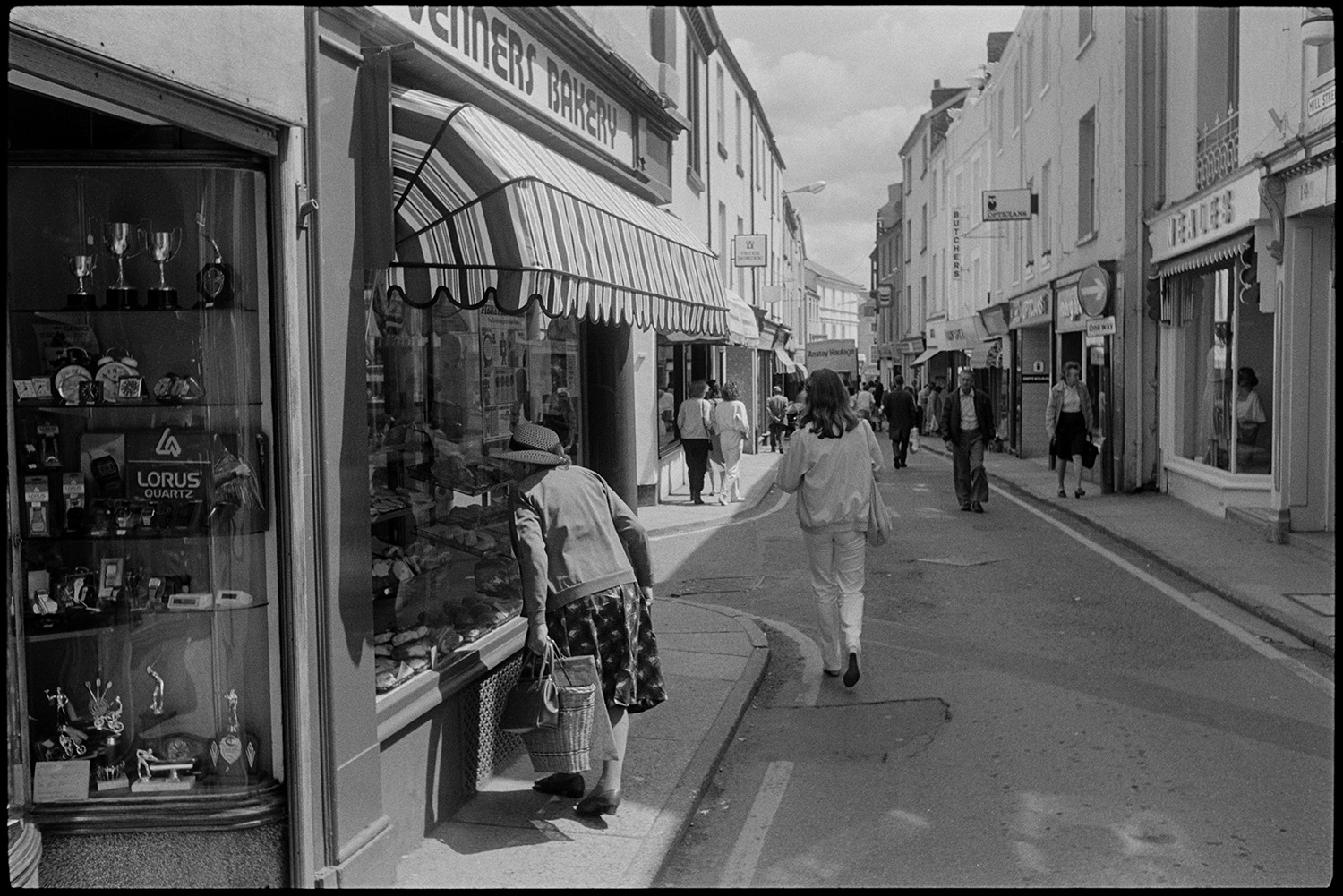 Street scenes with woman looking into shop window. 
[A woman looking into the shop front window of a bakery in Mill Street, Bideford. Other shoppers and passers by can be seen along the street, as well as shop fronts, including Heales.]