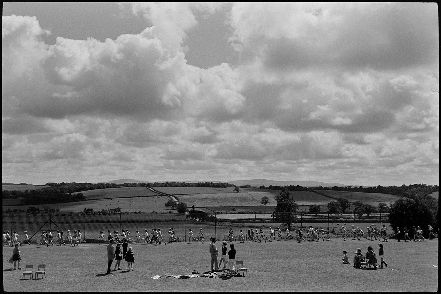 School sports day, children, races and spectators. 
[Children taking part in a long distance run around the school field at Winkleigh School at a sports day where children from Black Torrington School, Northlew School, Dolton School and Highampton School were competing. Other children and teachers are watching them.]