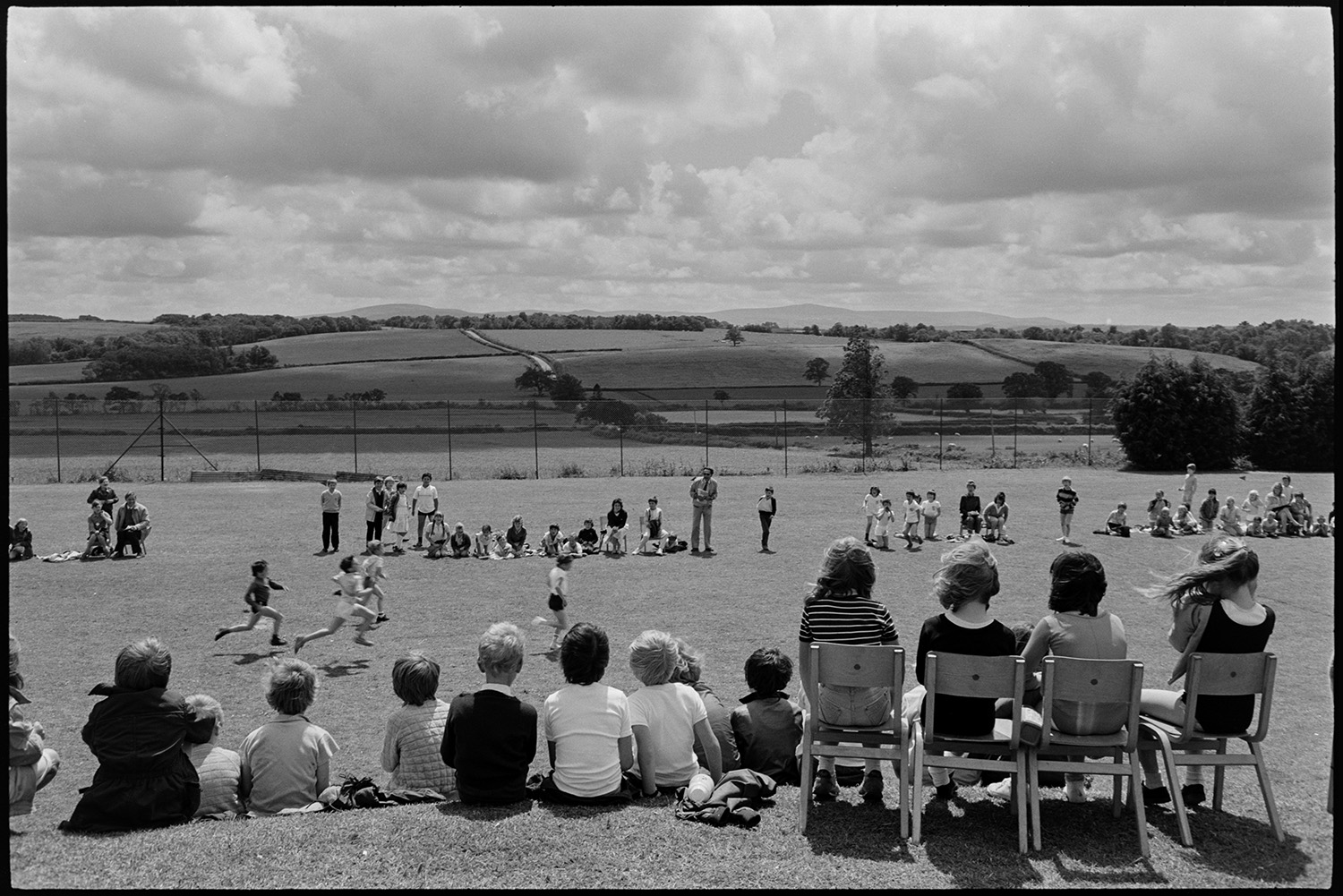 School sports day, children, races and spectators. 
[Children taking part in a running race on the school field at Winkleigh School at a sports day where children from Black Torrington School, Northlew School, Dolton School and Highampton School were competing. Other children and teachers are watching them from the side of the track. Some of the children in the foreground are sat on chairs.]