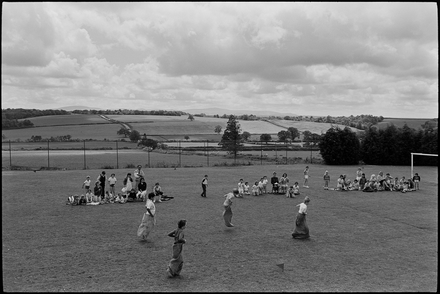 School sports day, children, races and spectators. 
[Children taking part in a sack race on the school field at Winkleigh School at a sports day where children from Black Torrington School, Northlew School, Dolton School and Highampton School were competing. Other children and teachers are watching them from the side of the track. Fields and trees can be seen in the background.]