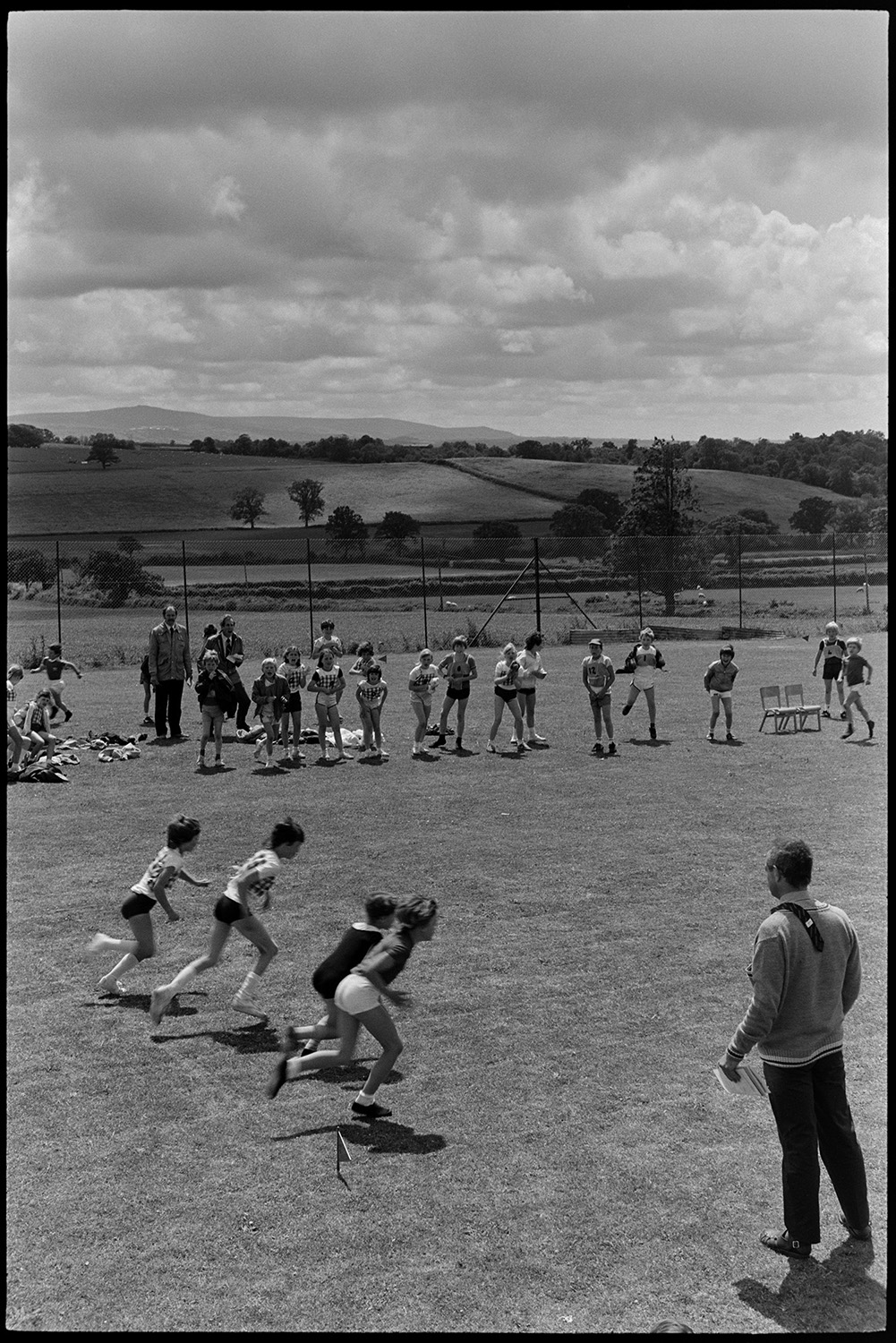 School sports day, children, races and spectators. 
[Children taking part in a running race on the school field at Winkleigh School at a sports day where children from Black Torrington School, Northlew School, Dolton School and Highampton School were competing. Other children are cheering from the side of the track.]
