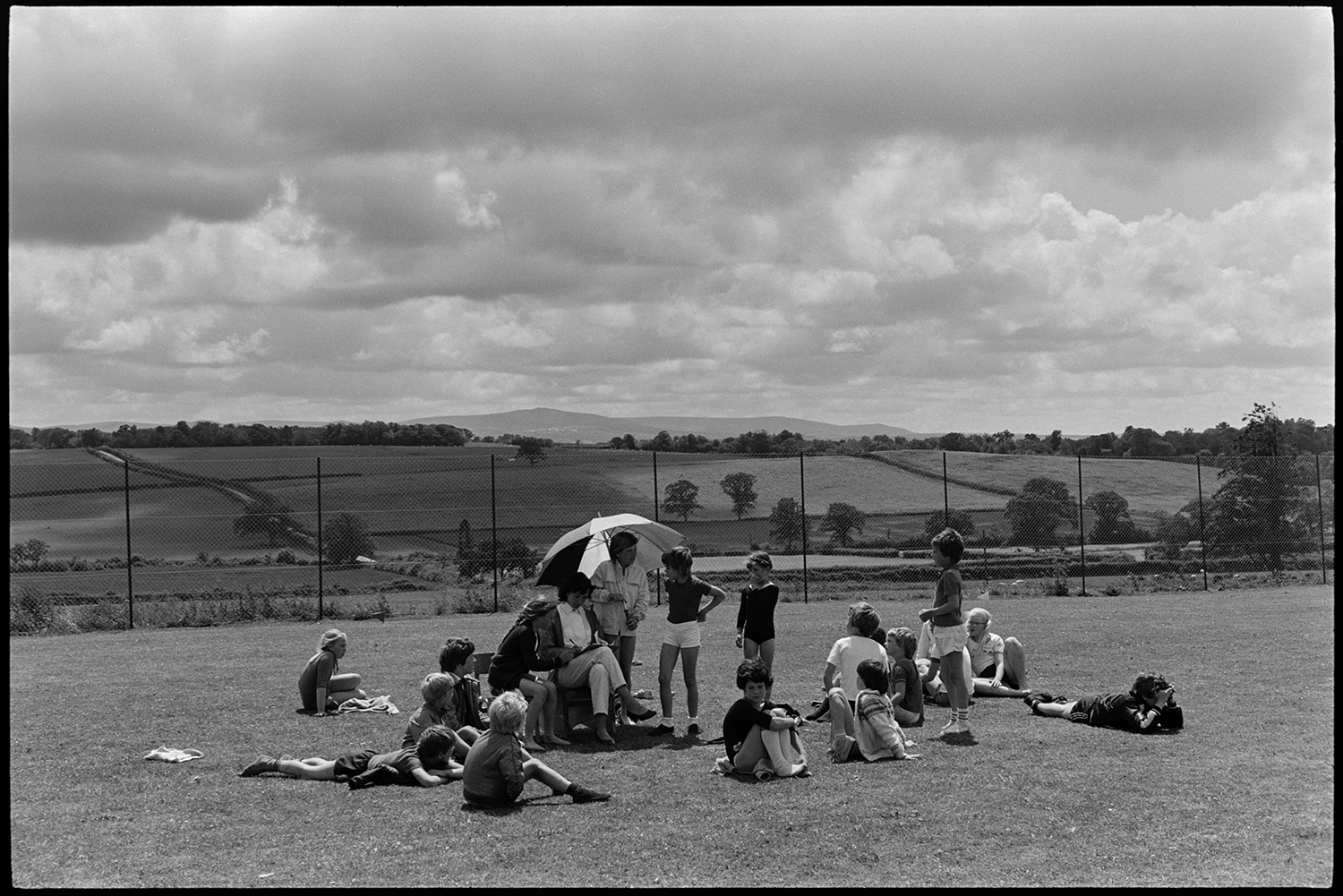 School sports day, children, races and spectators. 
[Children sat on the school field at Winkleigh School resting at a sports day where children from Black Torrington School, Northlew School, Dolton School and Highampton School were competing. A teacher is making notes and a child is holding an umbrella to shield her from the sun. A landscape of fields and trees can be seen in the background.]