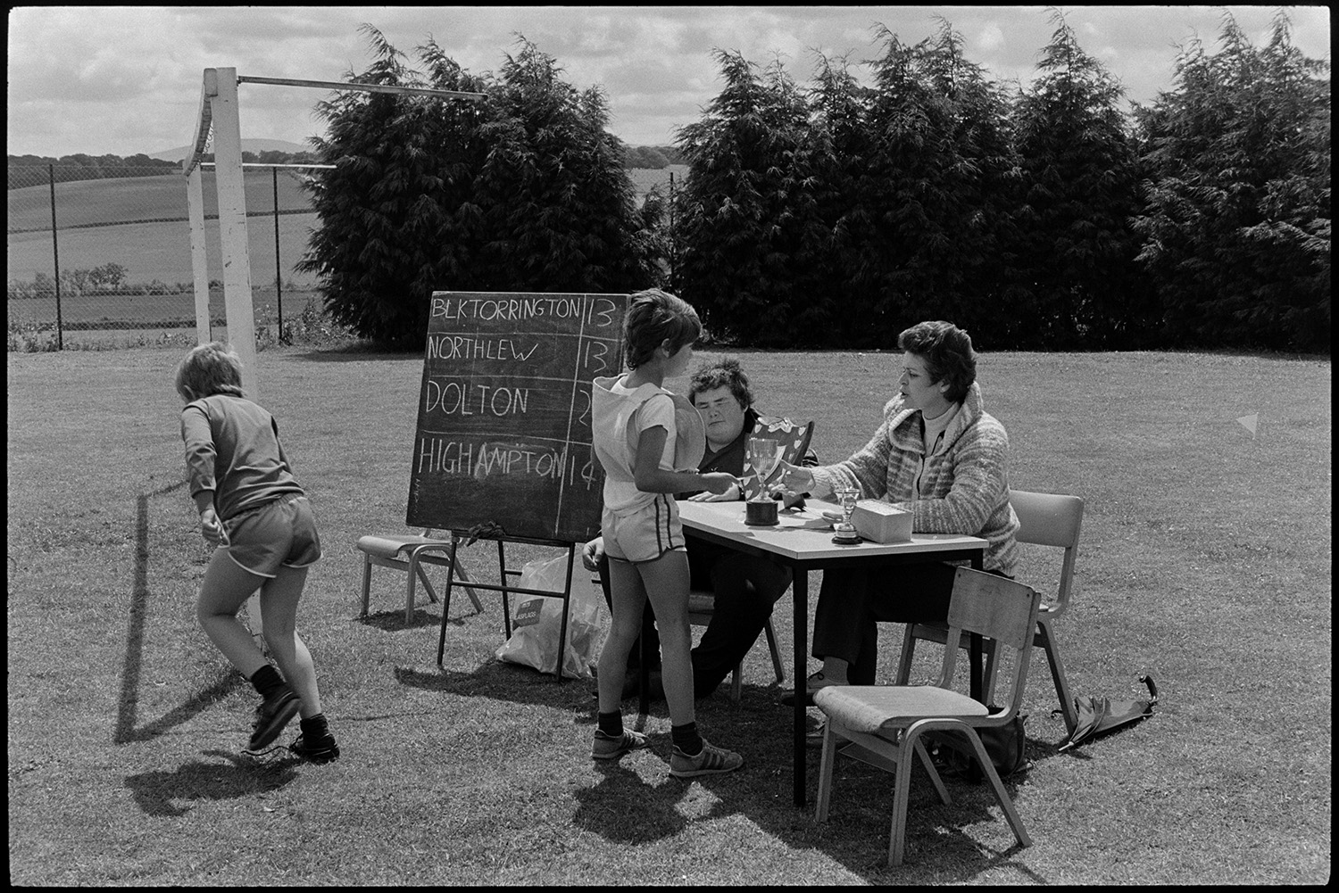 School sports day, children races, prize giving, cups. 
[Children talking to a teacher sat at a table with trophies on Winkleigh school sports field at a sports day where children from Black Torrington School, Northlew School, Dolton School and Highampton School were competing. The schools are listed on a chalkboard next to the table, with the number of points they have won.]