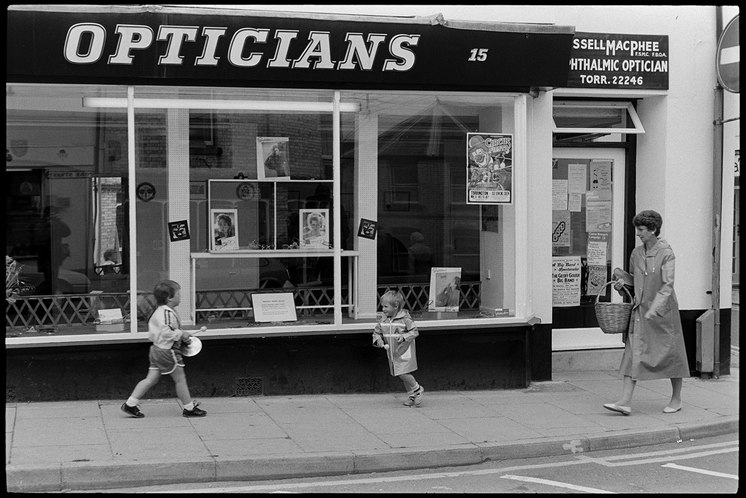 Street scenes with passers by and people chatting, woman postal worker. 
[A woman and two children walking past the shop front window of an Opticians in Torrington. The woman is carrying a basket.]