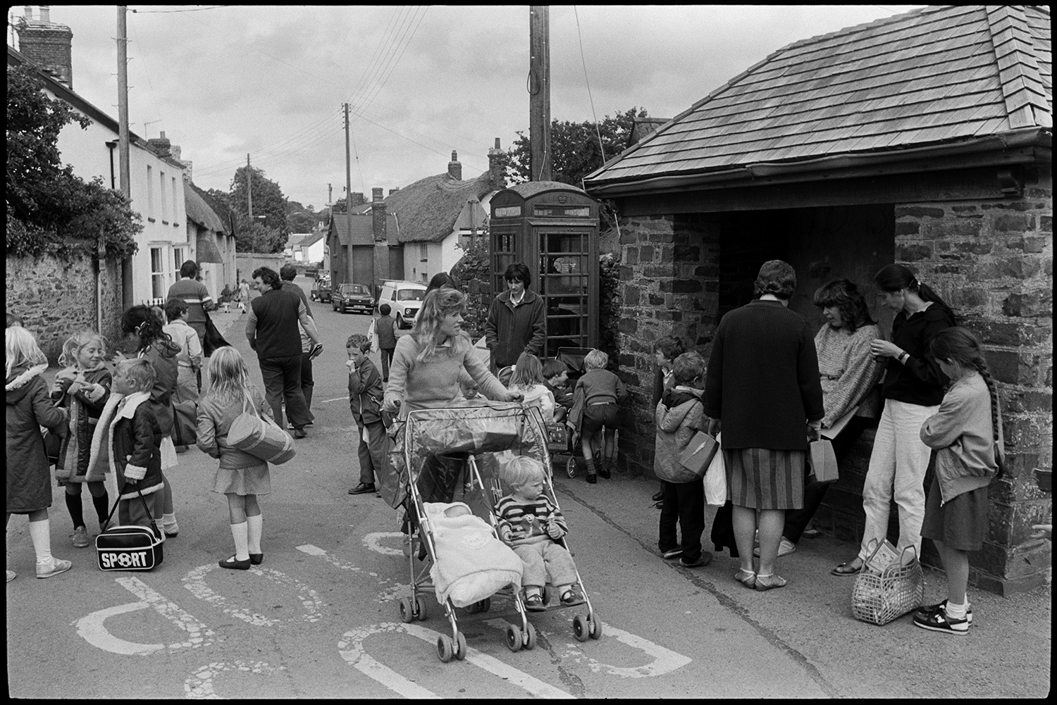 Women, mothers and children after school, pushchair. 
[Mothers greeting children after school outside a bus shelter and telephone box in Dolton. A woman in the foreground is pushing a double pram.]