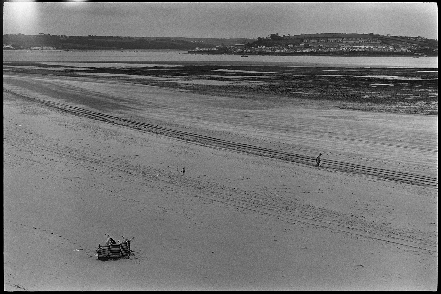 View across estuary with beach and distant power station. 
[People on the beach at Braunton Burrows. A windbreak is also visible with a pram in the foreground. Instow (?) can be seen across the estuary.]