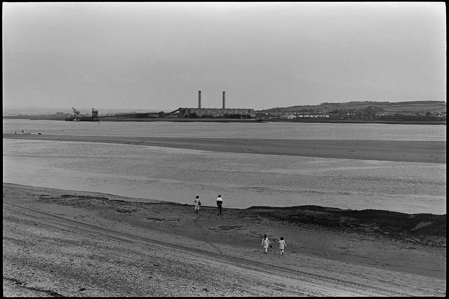 View across estuary with beach and distant power station. 
[A woman, man and two children walking along the beach at Braunton Burrows. They are looking across the estuary to Yelland Power Station.]
