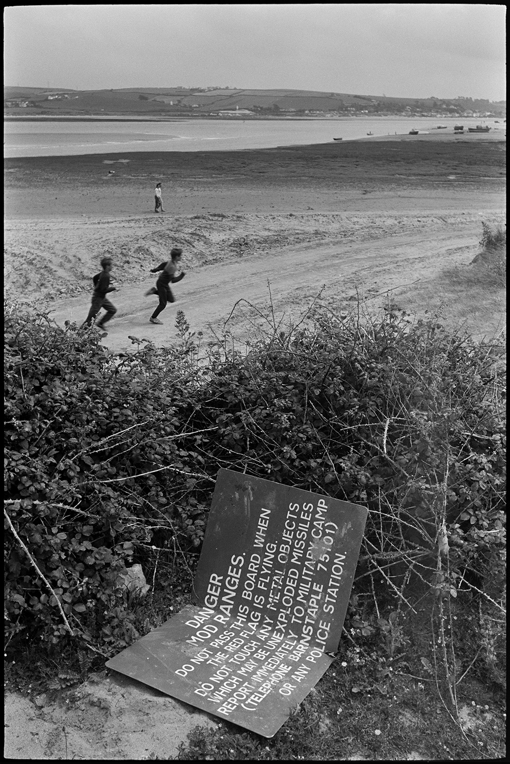 View across estuary with beach and distant power station. 
[Children playing on the beach at Braunton Burrows. A sign in foliage in the foreground is warning about metal objects and unexploded missiles. The estuary is visible in the background.]
