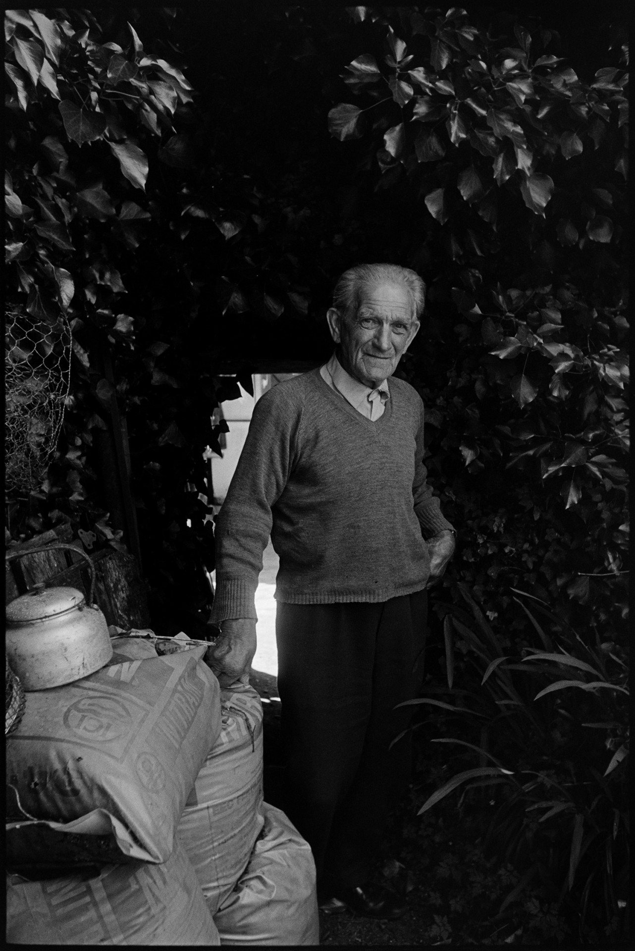 Man chatting in garden, standing in doorway. 
[Bill Hutchins standing by a pile of sacks and an archway or gateway surrounded by foliage, in his garden at Dolton.]