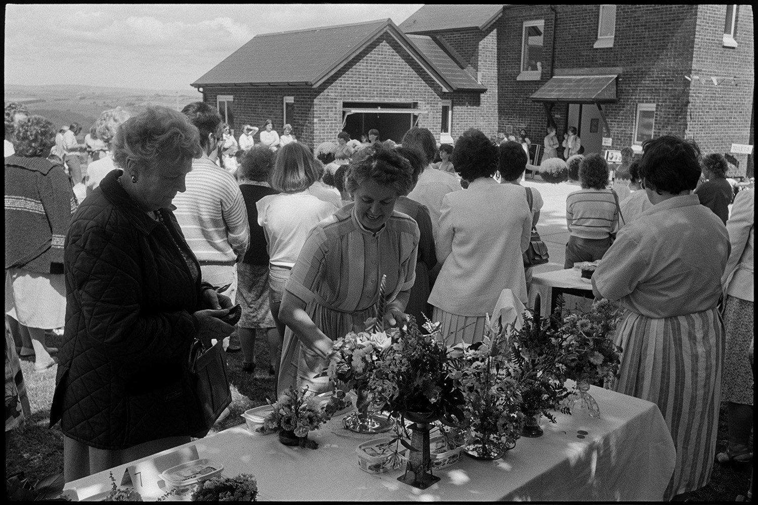 Stalls at fete, skittle alley cakes, raffle stall, pony ride. <br />
[Women looking at flower displays on a stall at High Bickington church fete held at the vicarage. People are gathered around watching majorettes performing in the background.]