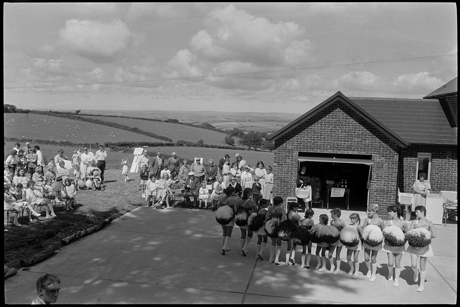 Majorettes at vicarage fete with cloudy landscape. <br />
[The Bluebirds Majorettes performing with pom poms at High Bickington church fete held at the vicarage. People are gathered around to watch them. Raffle tickets are being sold in the open garage.[