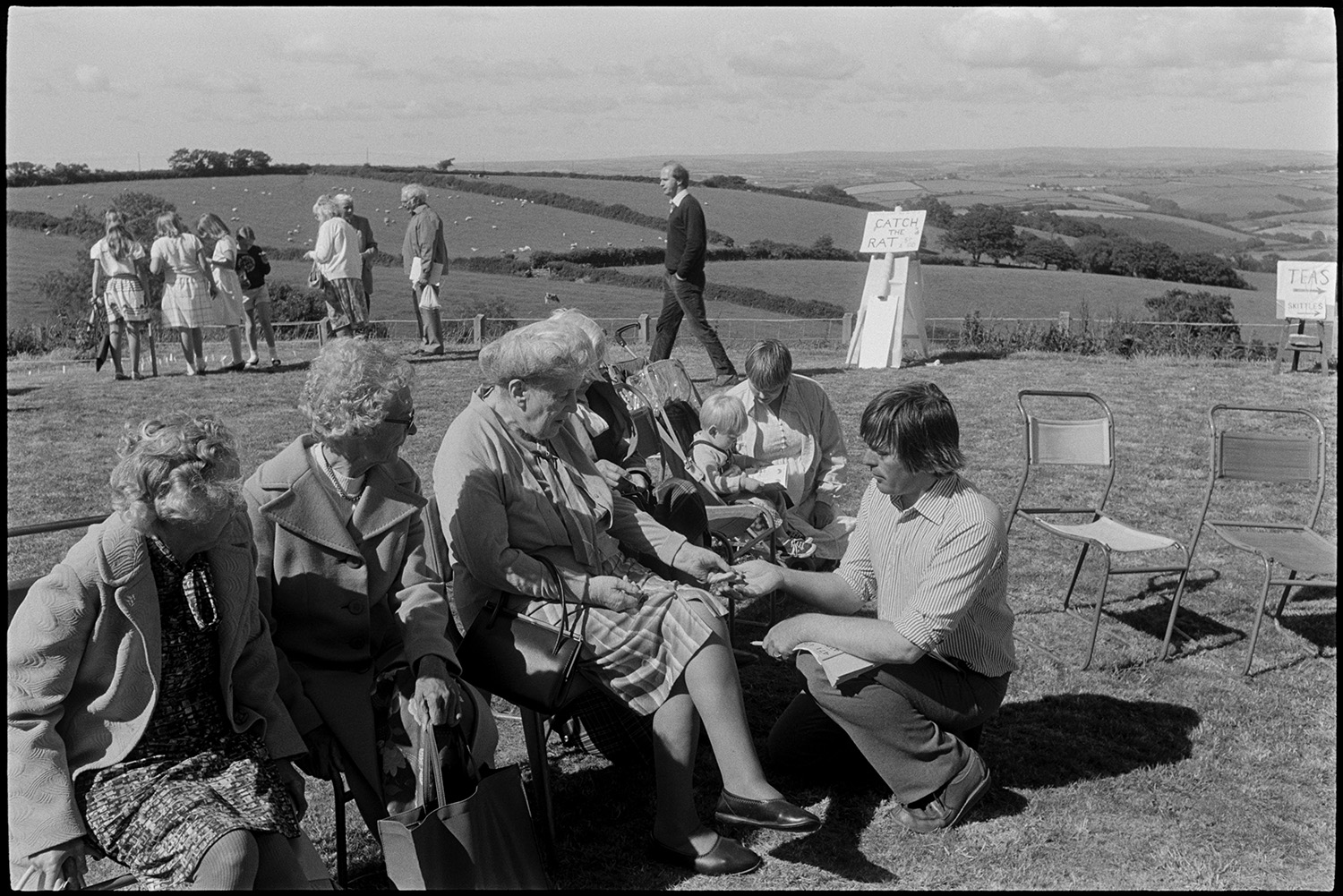 Man selling raffle tickets to elderly women at fete. 
[A man selling raffle tickets to women, including Mary Pickard and Mrs Piddler, at Atherington church fete held at the vicarage. The women are sat on chairs on the grass. A 'catch the rat' game can be seen in the background.]