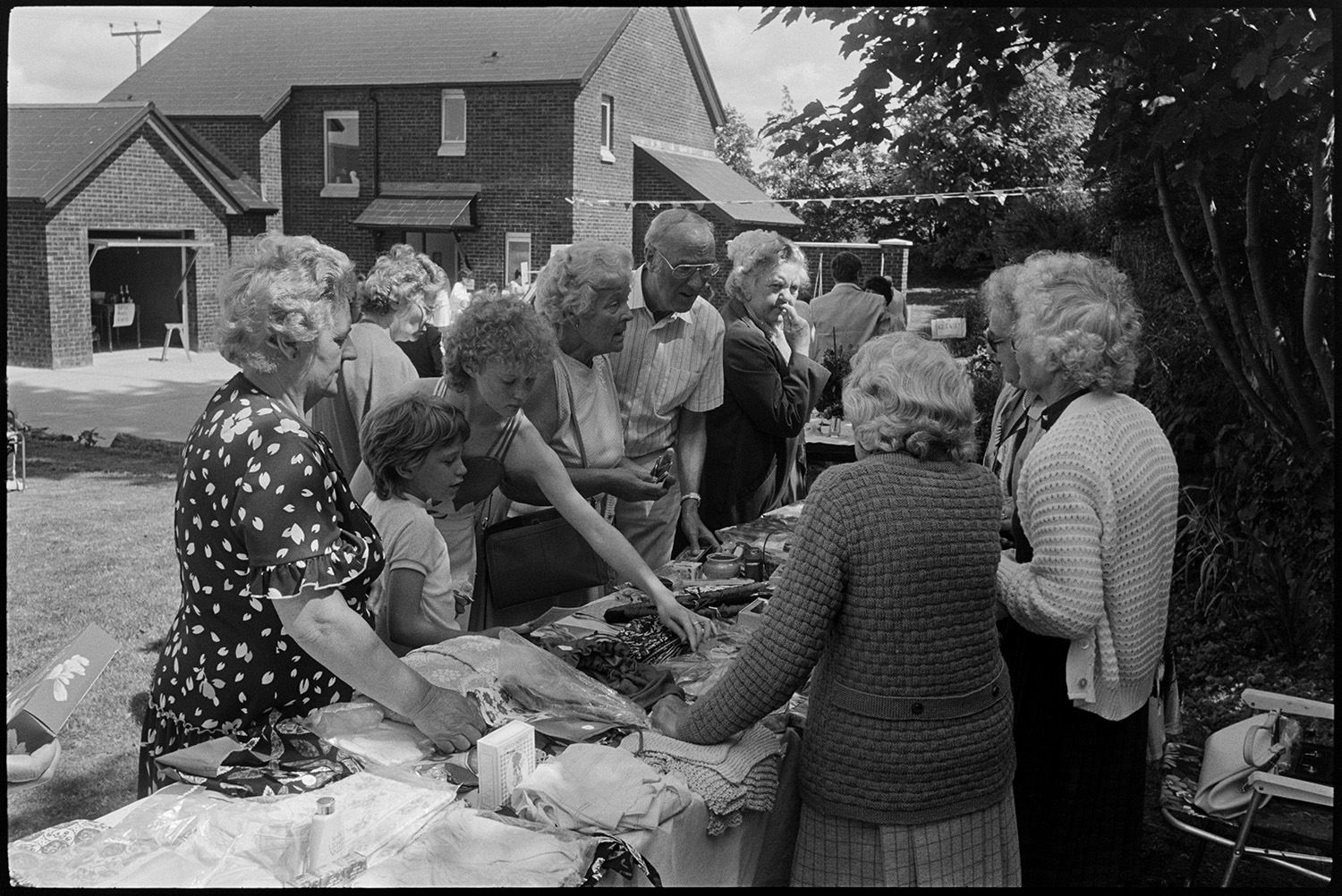 Fete stalls, games, majorettes. 
[Men, women and children looking at a jumble stall at Atherington church fete held at the vicarage. They are talking to the women running the stall.]