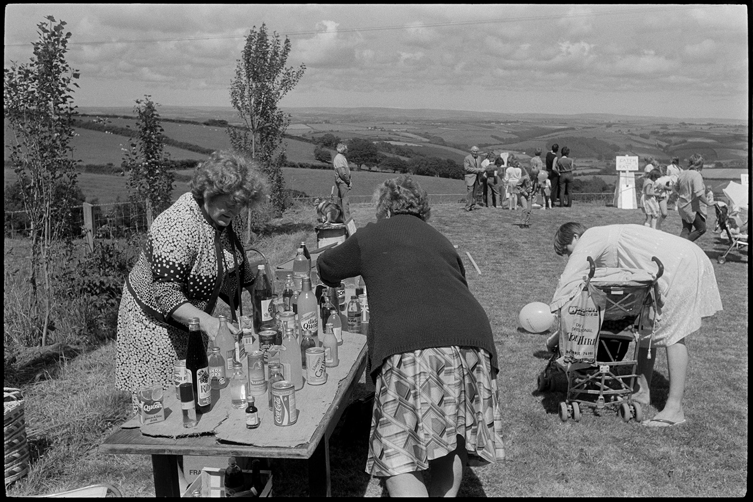 Fete stalls, games, majorettes. 
[Women organising the bottle stall at Atherington church fete held at the vicarage. A women is tending to a child in a pram next to the stall. Other games and stalls can be seen in the background.]