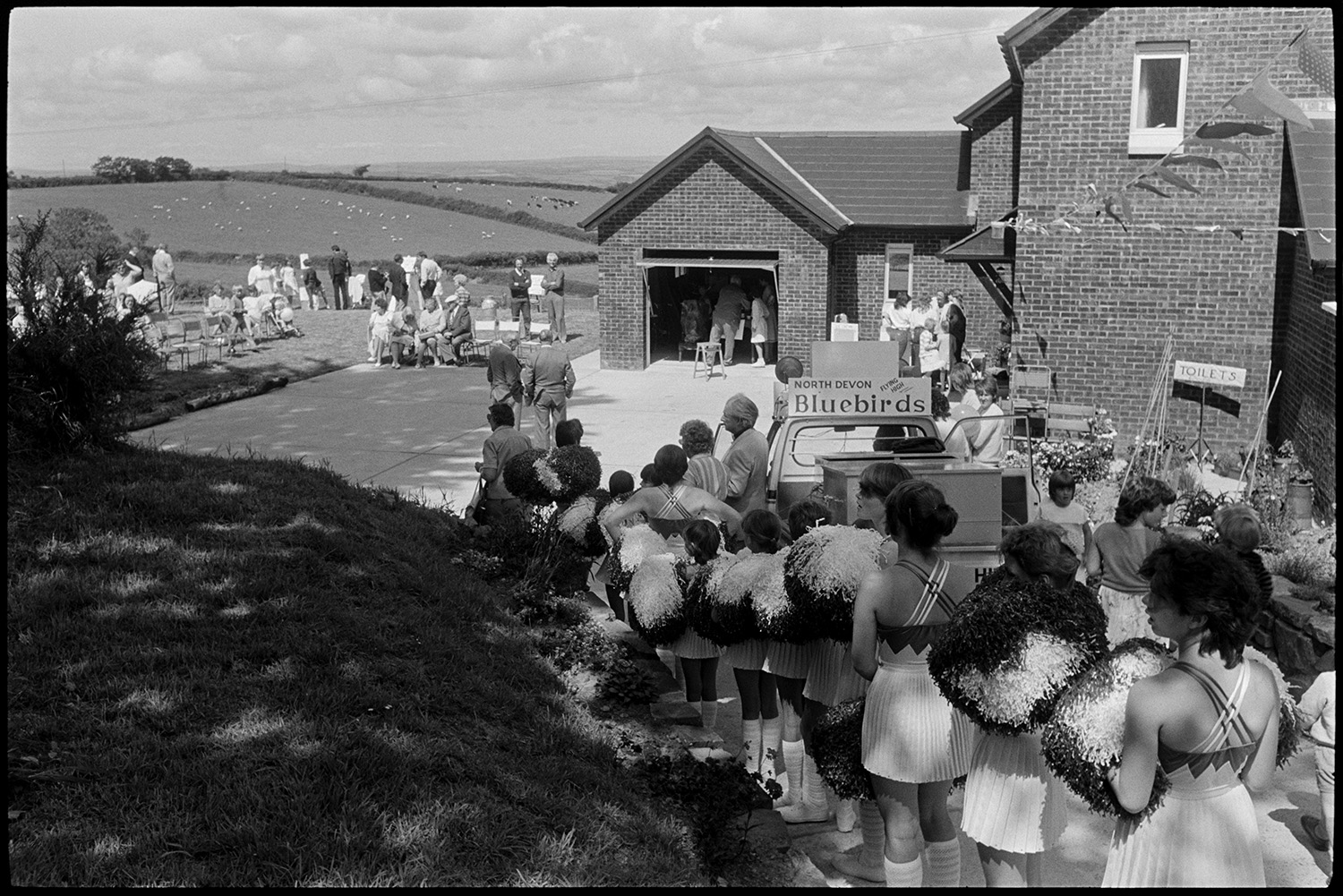 Fete stalls, games, majorettes. 
[The Bluebirds Majorettes getting ready to perform, with pom poms, at Atherington church fete held at the vicarage. People are gathered around outside the house to watch the performance.]
