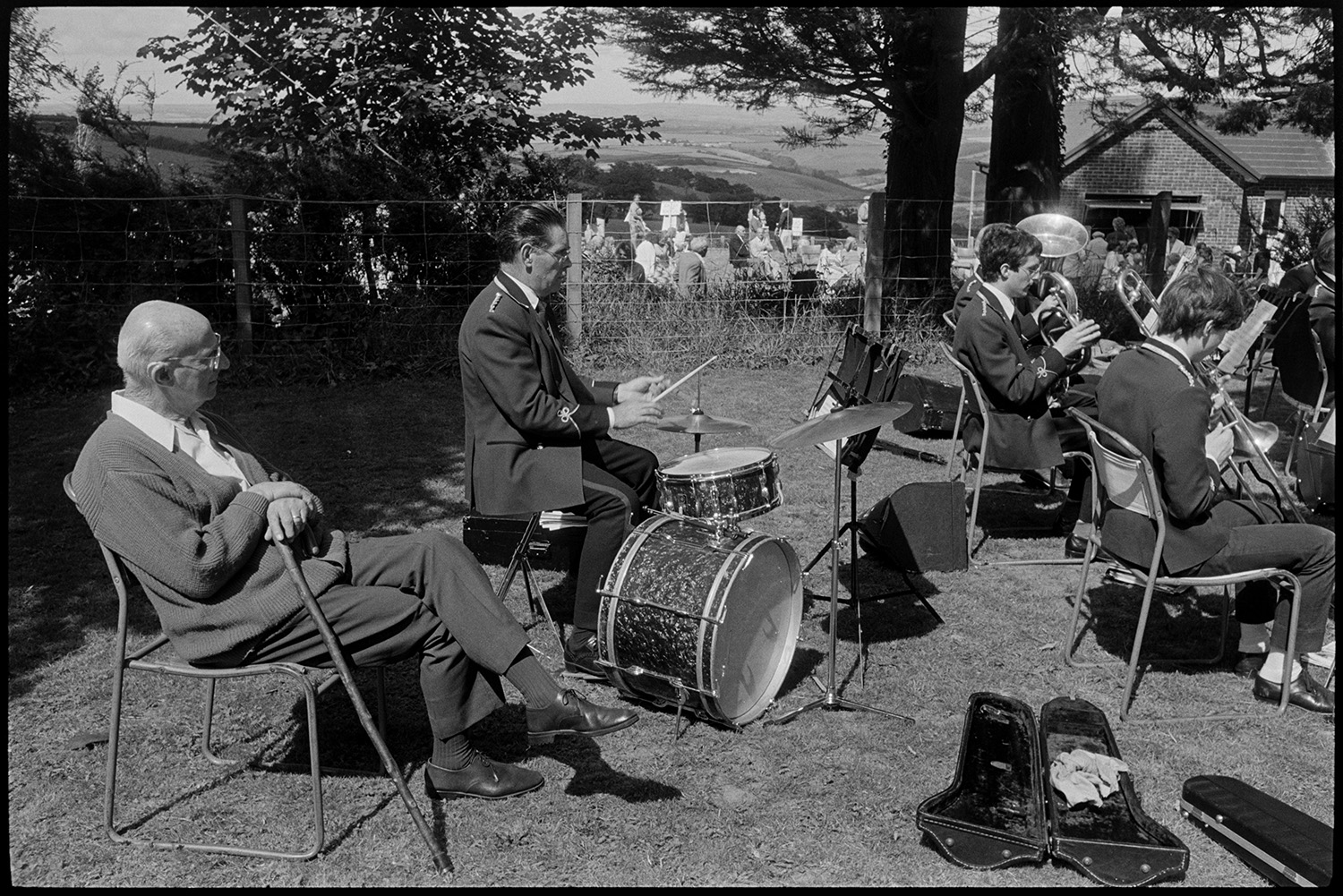 Fete, spectators, Brass Band, sideshows, games, pony rides. 
[A man sat on a chair listening to a band at Atherington church fete. He is sat by a man playing a drum kit.  They are playing in a field by a tree. People looking at stalls in the vicarage garden can be seen in the background.]