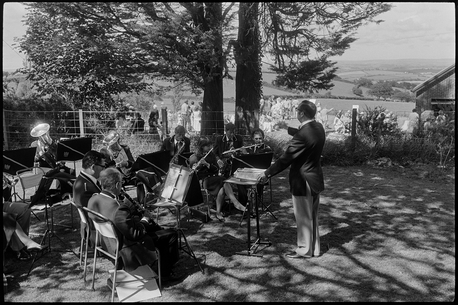 Fete, spectators, Brass Band, sideshows, games, pony rides. 
[A man conducting a band at Atherington church fete held at the vicarage. They are playing a field by a tree. People looking at stalls in the vicarage garden can be seen in the background.]