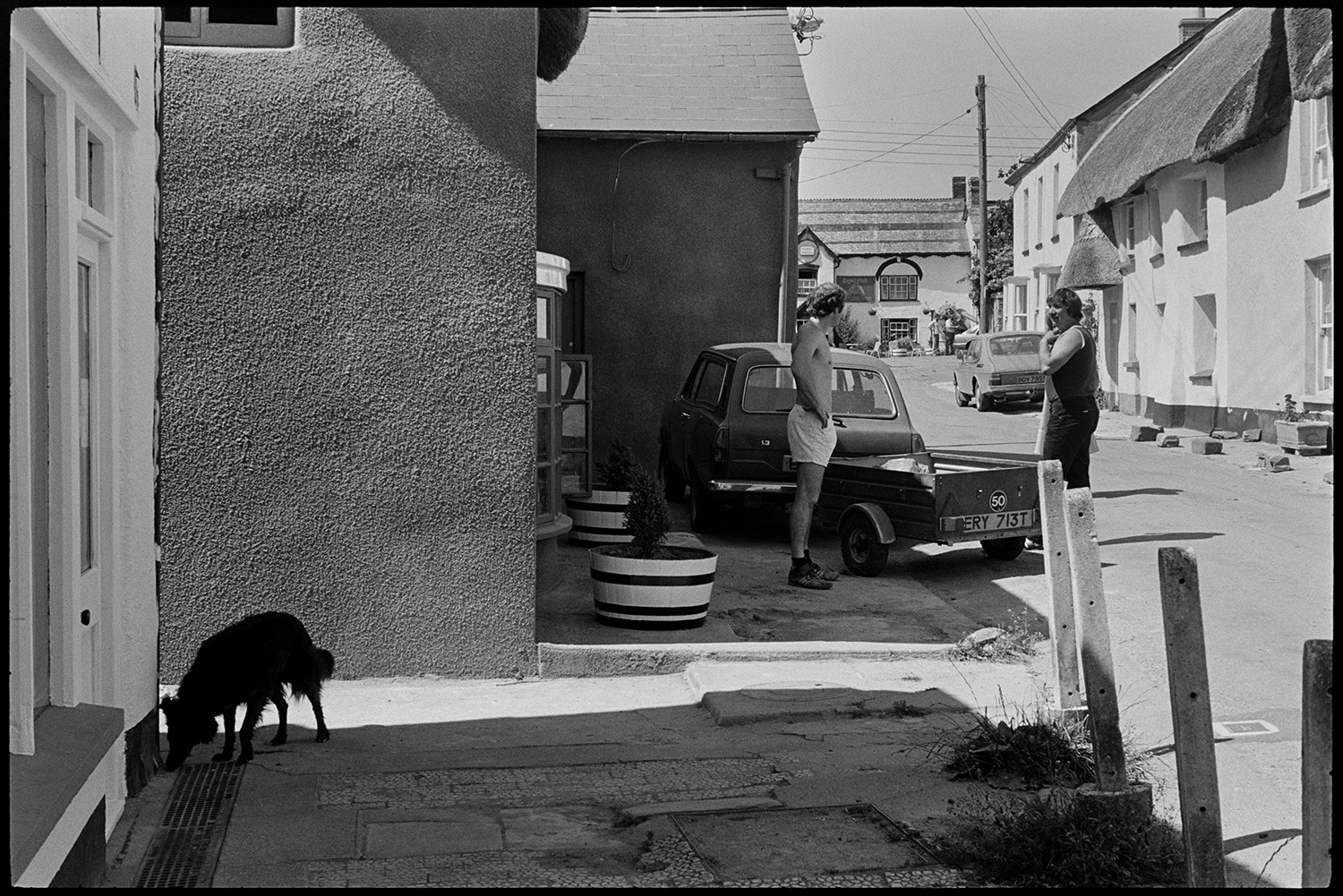 Street scenes, bicycles, people mending car.
[Two men talking by a car with a trailer parked in Fore Street, Dolton. A dog is in the foreground and thatched cottages and the Royal Oak pub are visible in the background.]