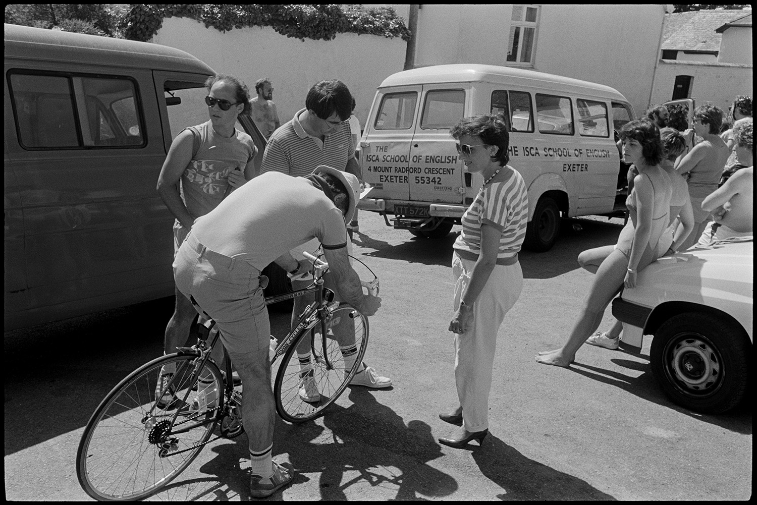 Street scenes, bicycles, people mending car.
[People gathered in Fore Street, Dolton for a bicycle event. A person is on a bicycle in the foreground. A minibus from the ISCA School of English is parked in the background.]