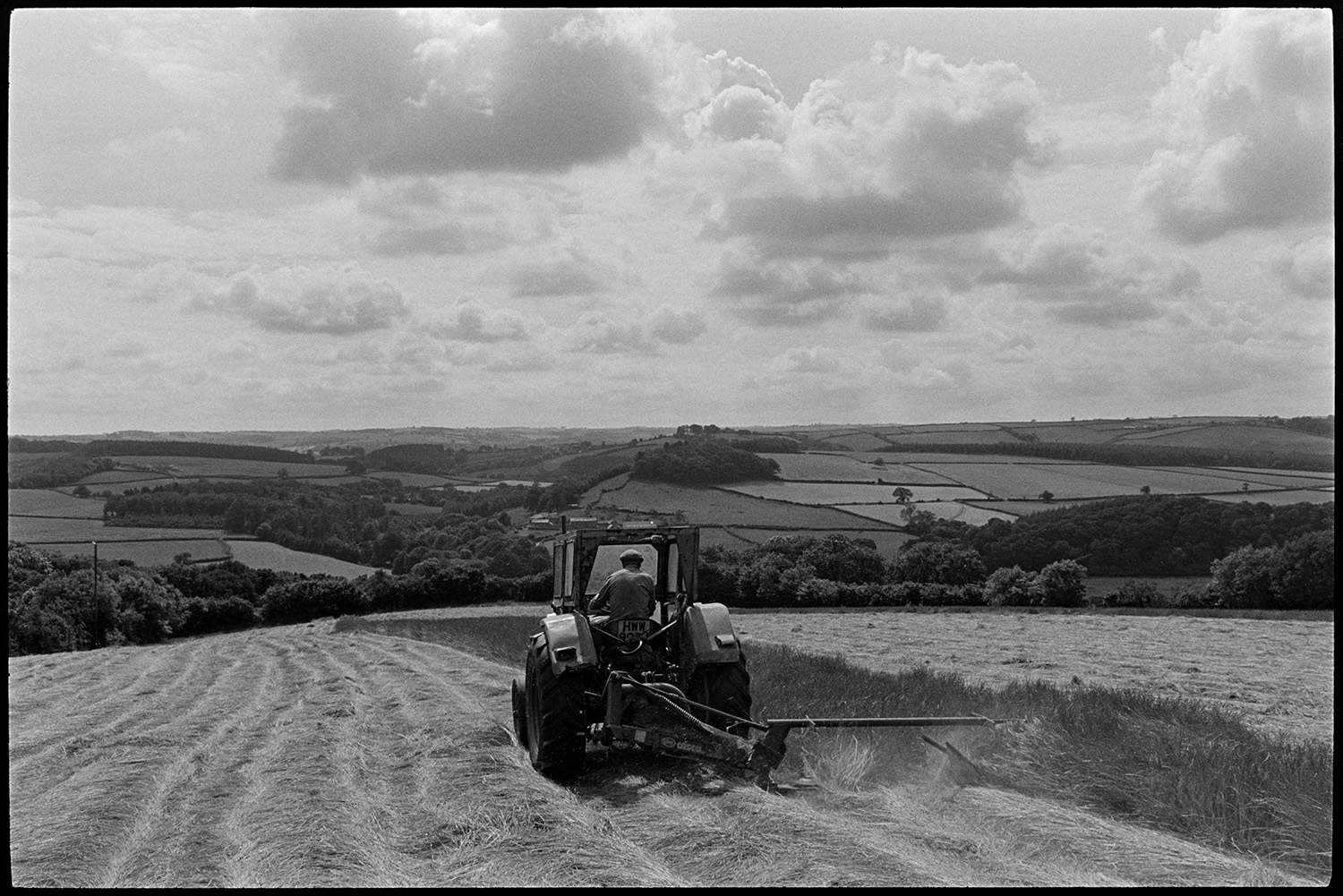 Tractor and grass cutter, mower in sunny landscape. 
[George Ayre using a tractor a mower to cut grass for hay, in a sunny field at Ashwell, Dolton. The tractor is moving away from the camera. Clouds are visible in the sky and a landscape of fields, hedgerows and trees can be seen in the background.]