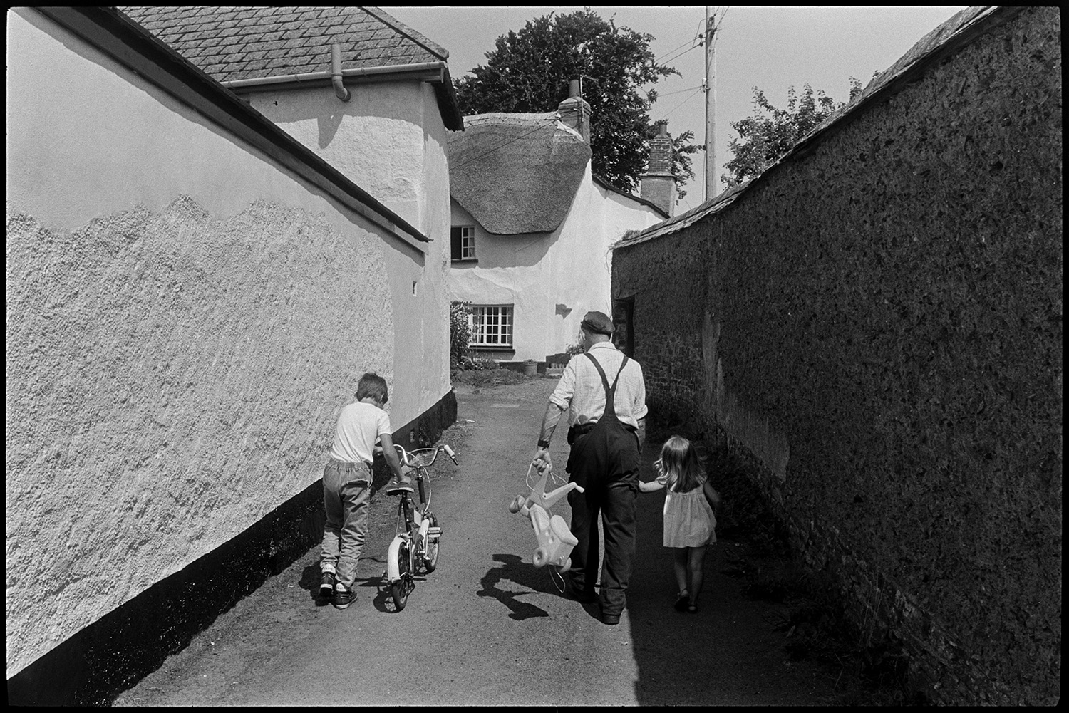 Street scenes. Man and children. 
[A man and two children walking along North Street, Dolton towards a thatched cottage. A girl is holding the man's hand while he carries her trike in his other hand, and a boy is pushing a bicycle.]