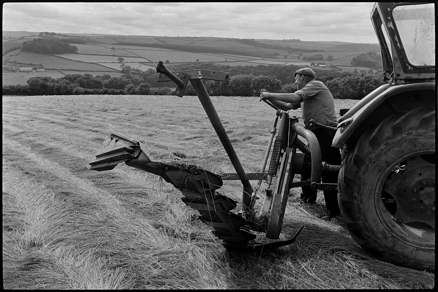 Tractor and grass cutter, mower, farmer folding up cutter. 
[George Ayre folding up a grass cutter attached to a tractor, in a field at Ashwell, Dolton. He has cut the field of grass for hay. A landscape of fields, hedgerows and trees is visible in the background.]