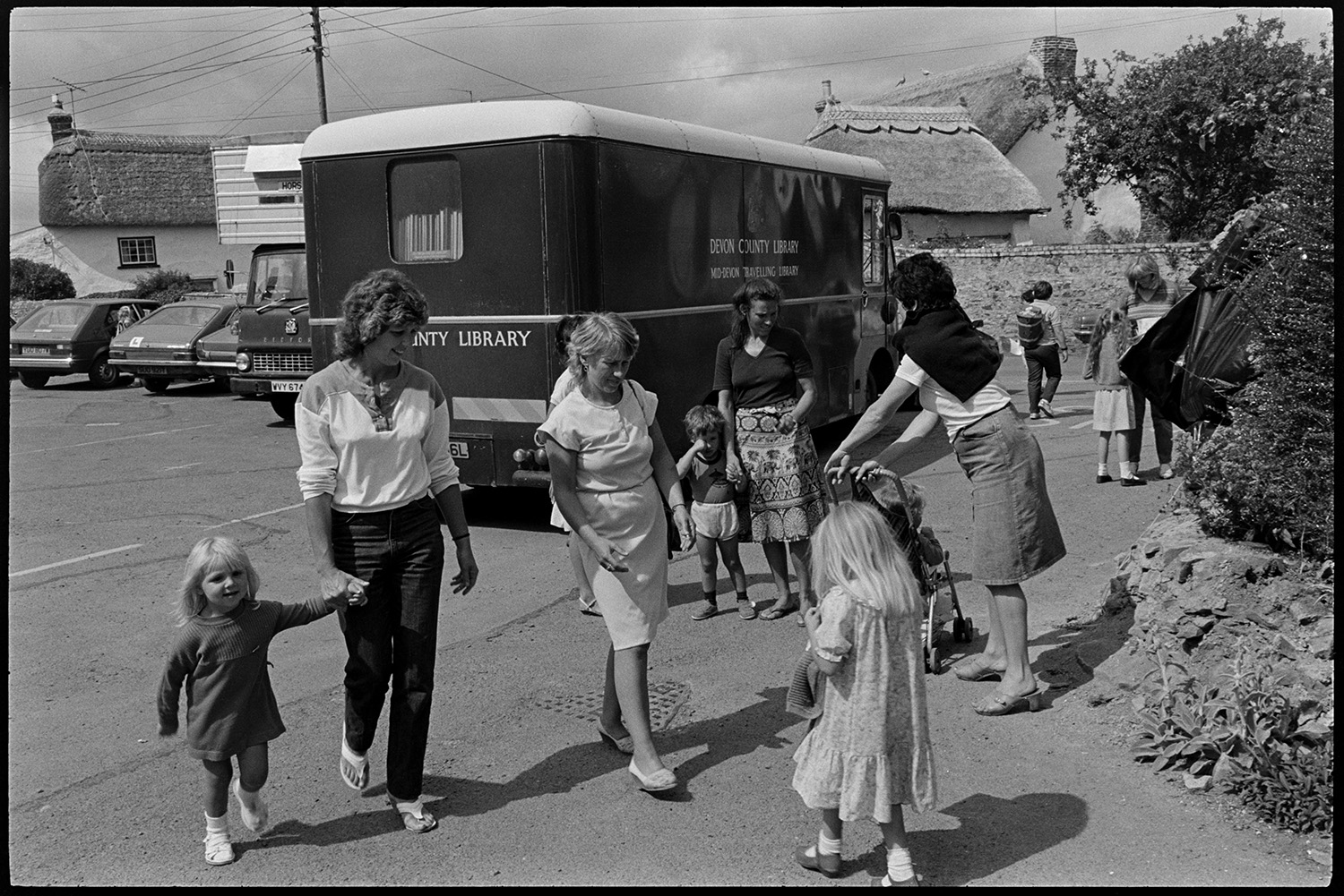 Library van parked in village, women and children after school. 
[Woman and children after school in a street in Dolton. They are stood by a library van. Thatched cottages and parked cars can be seen in the background.]