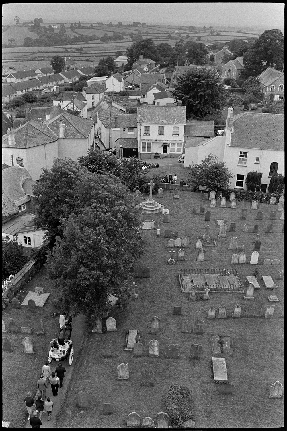 Procession in churchyard with Fair Queen at start of Fair. Graveyard and gravestones. 
[A view taken from Winkleigh Church tower of people processing down the church path before Winkleigh Fair. They are following the Winkleigh Fair Queen in a horse drawn carriage. Gravestones in the churchyard, the war memorial and houses in the village can be seen.]