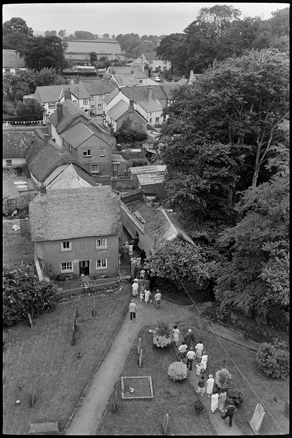 Procession in churchyard with Fair Queen at start of Fair. Graveyard and gravestones. 
[A view taken from Winkleigh Church tower of people processing down the church path before Winkleigh Fair. Gravestones in the churchyard and houses in the village can be seen.]