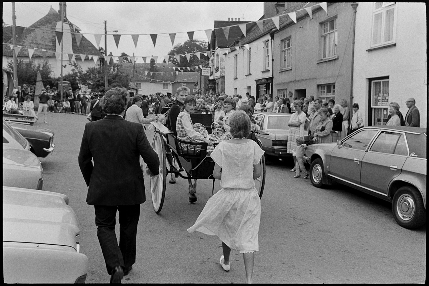 Fair Queen arriving in village in pony trap. 
[The Winkleigh Fair Queen arriving in Winkleigh Square for the start of the Fair in a horse drawn open carriage. A crowd has gathered to watch and the street is decorated with bunting.]