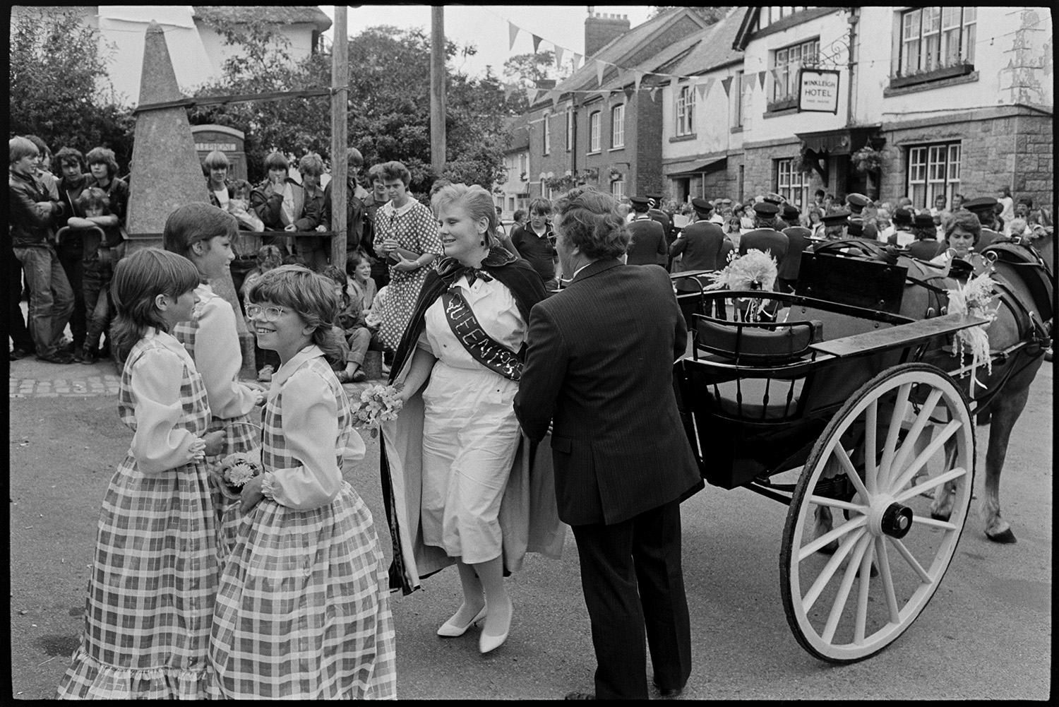 Fair Queen arriving in village in pony trap. 
[The Winkleigh Fair Queen arriving in Winkleigh Square for the start of the Fair in a horse drawn open carriage. She is getting out of the carriage with her attendants. A crowd of people are watching and the street is decorated with bunting. The Winkleigh Hotel is visible in the background.]