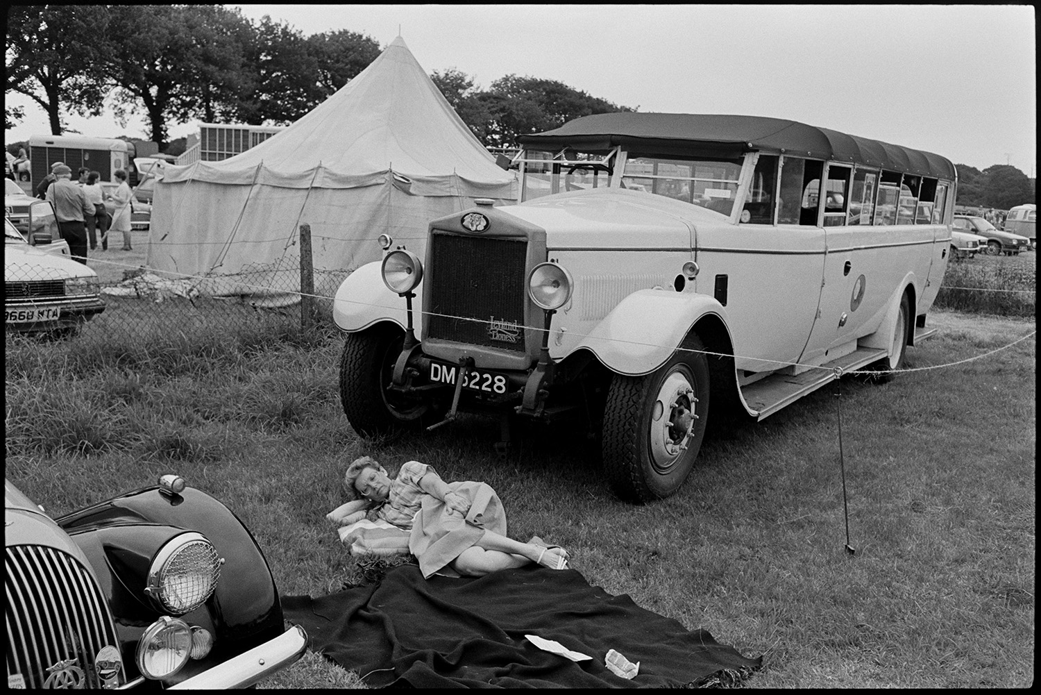 Gymkhana at Fair, horses, ponies and riders. Old bus. 
[A woman lying on a picnic rug in front of a charabanc  at Winkleigh Fair. A tent or small marquee can be seen in the background.]