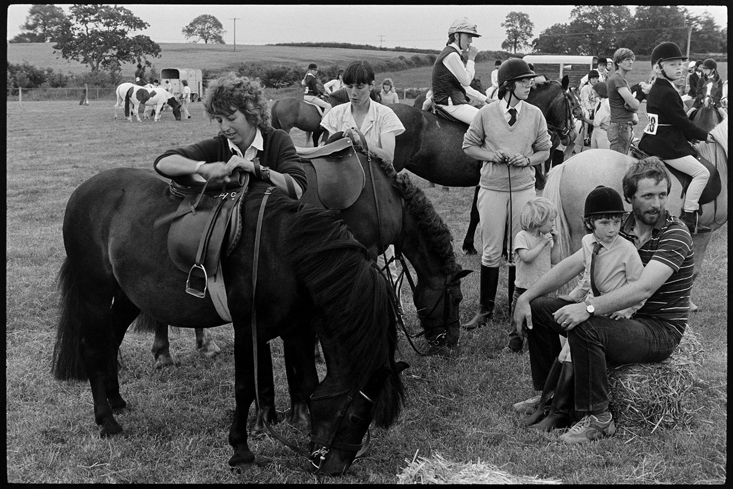 Gymkhana at Fair, horses, ponies and riders. Old bus. 
[Two women sorting the saddles on horses at a gymkhana at Winkleigh Fair. Riders, horses and spectators are gathered in the field in the background.]