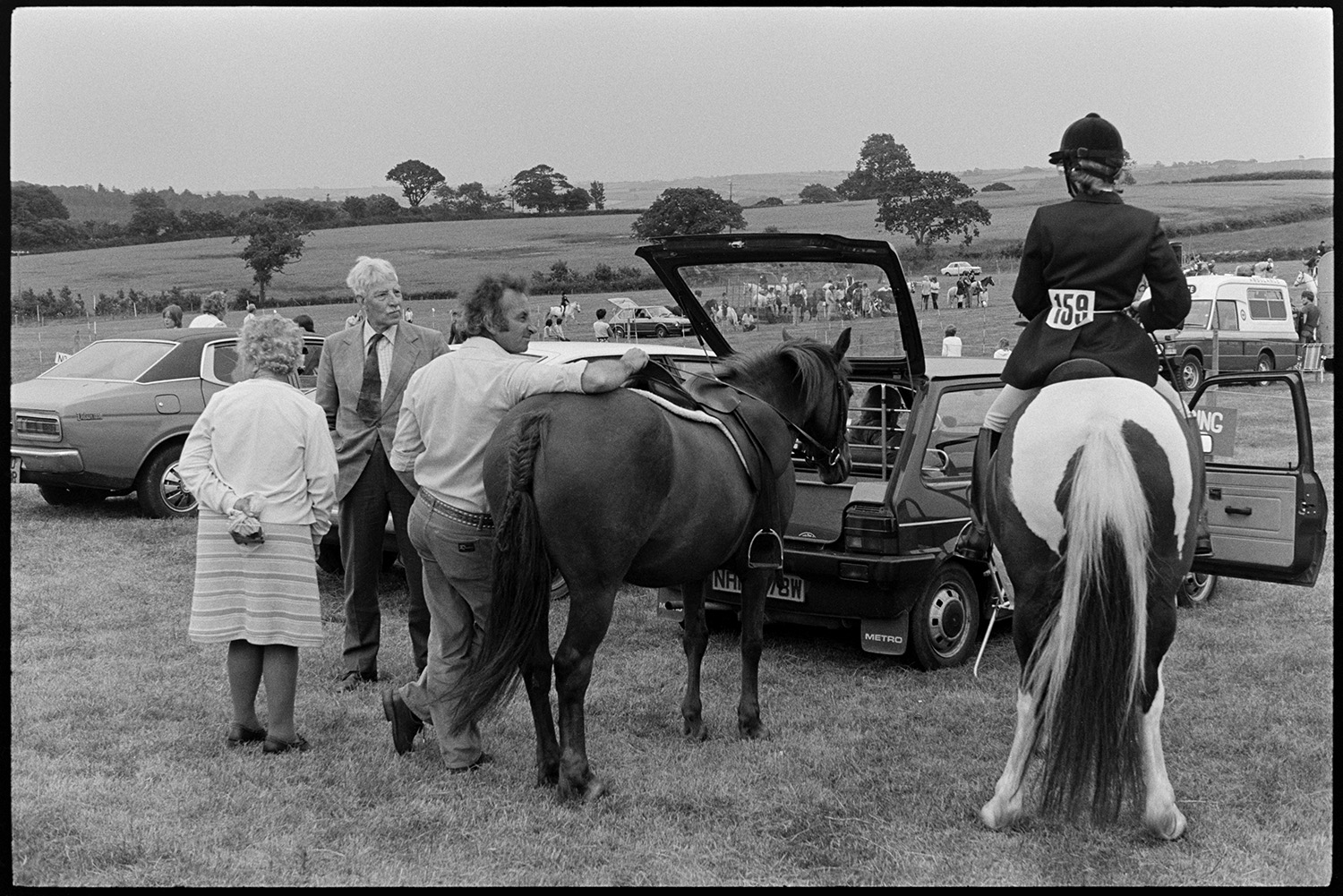 Gymkhana at Fair, horses, ponies and riders. Old bus. 
[Men, a woman and two horses gathered by a car in a field at a gymkhana at Winkleigh Fair. A rider is sat on one of the horses. Other people at the gymkhana can be seen in the background.]