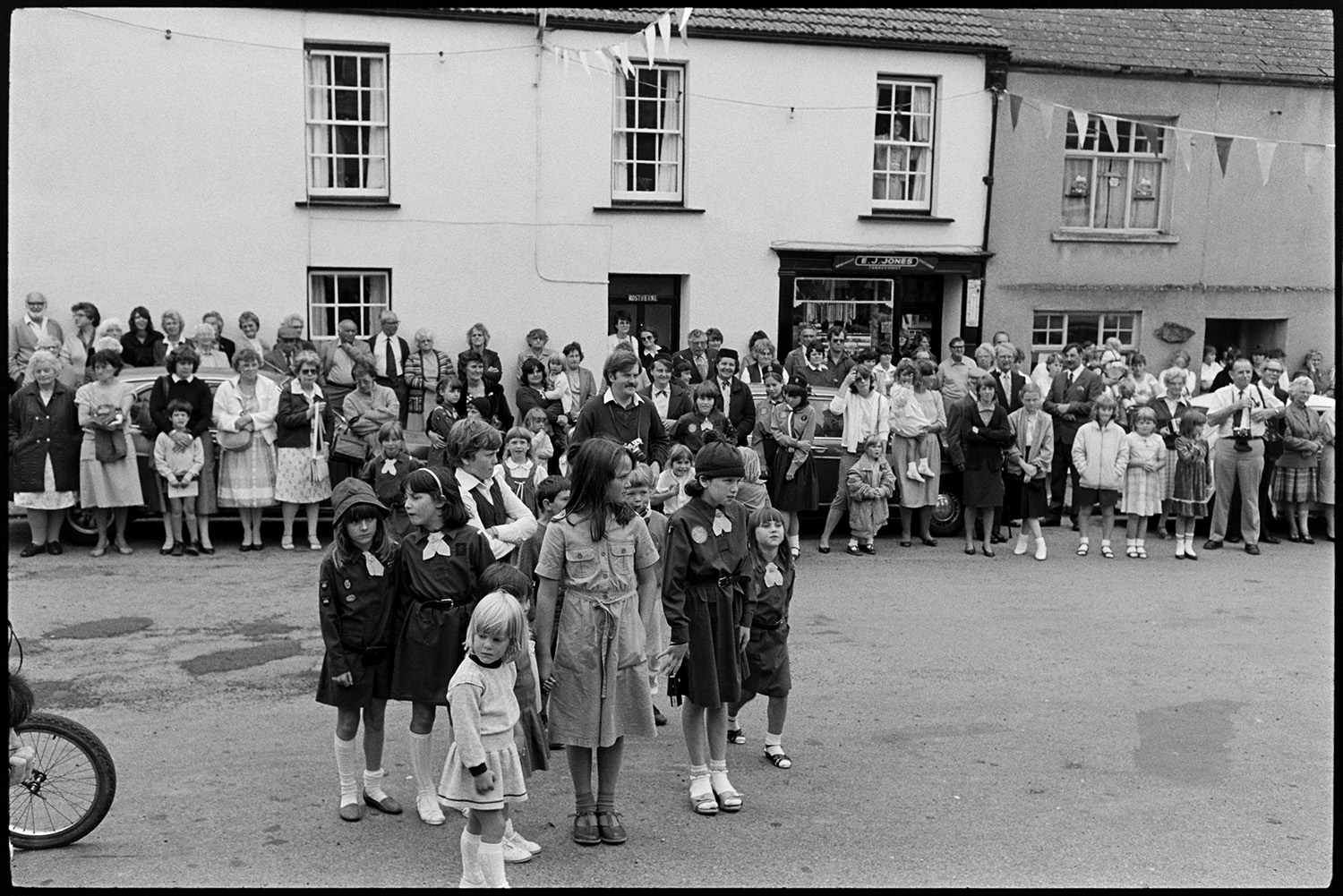 Fair Queen and attendants in village square, spectators waiting around. Photographer. 
[A group of Brownies in Winkleigh village square at Winkleigh Fair. A crowd of people are watching in the background and the street is decorated with bunting.]