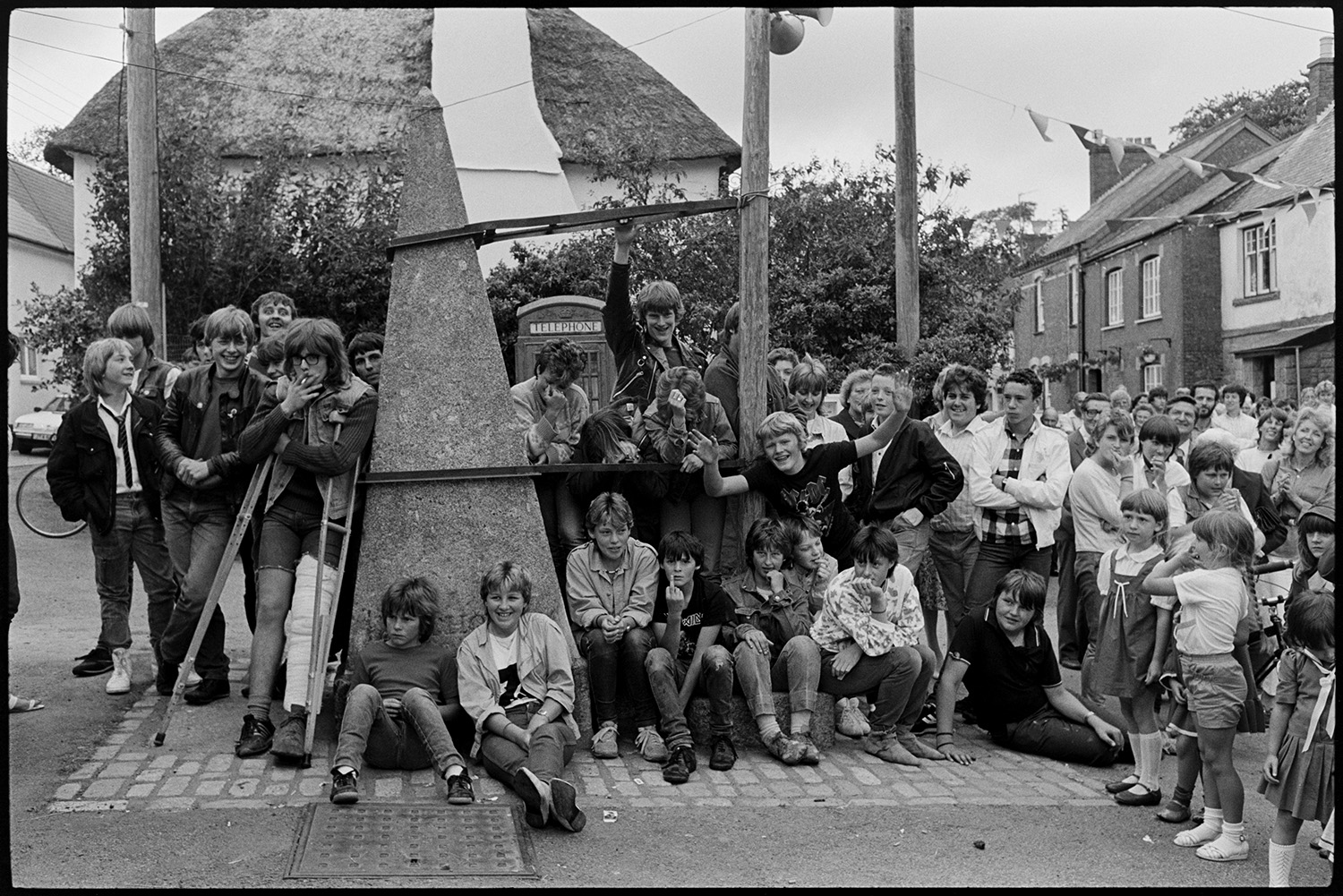 Fair Queen and attendants in village square, spectators waiting around. Photographer. 
[Children and teenagers gathered around the village pump in Winkleigh square waiting for the start of Winkleigh Fair. One teenager has a pair of crutches and is smoking.]