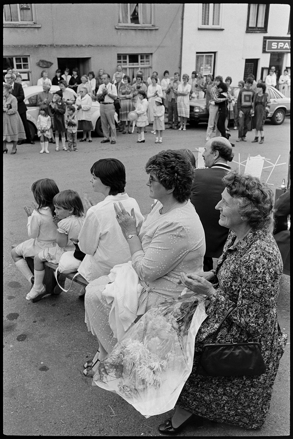 Fair Queen and attendants in village square, spectators waiting around. Photographer. 
[Women and children sat on a bench watching entertainment in Winkleigh village square at Winkleigh Fair. One of the women has a bouquet of flowers on her lap. Other people are watching on the other side of the street, by the Spar shop.]