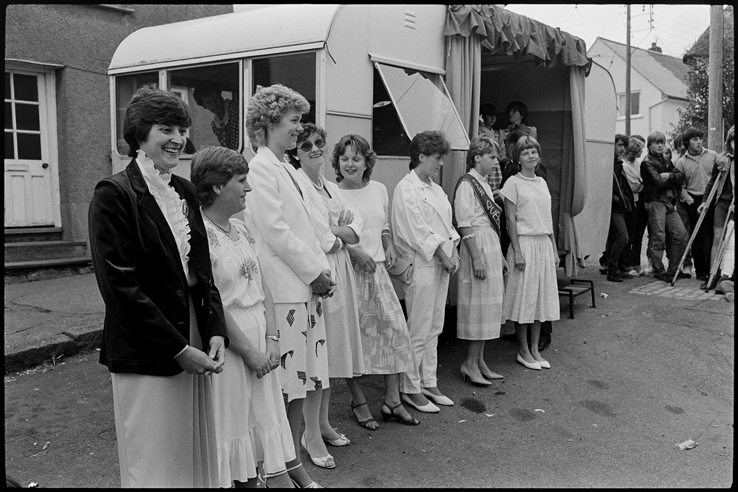 Fair Queen and attendants in village square, spectators waiting around. Photographer. 
[The Winkleigh Fair Queen, attendants and women stood outside a caravan in Winkleigh square which has been made into a stage.]